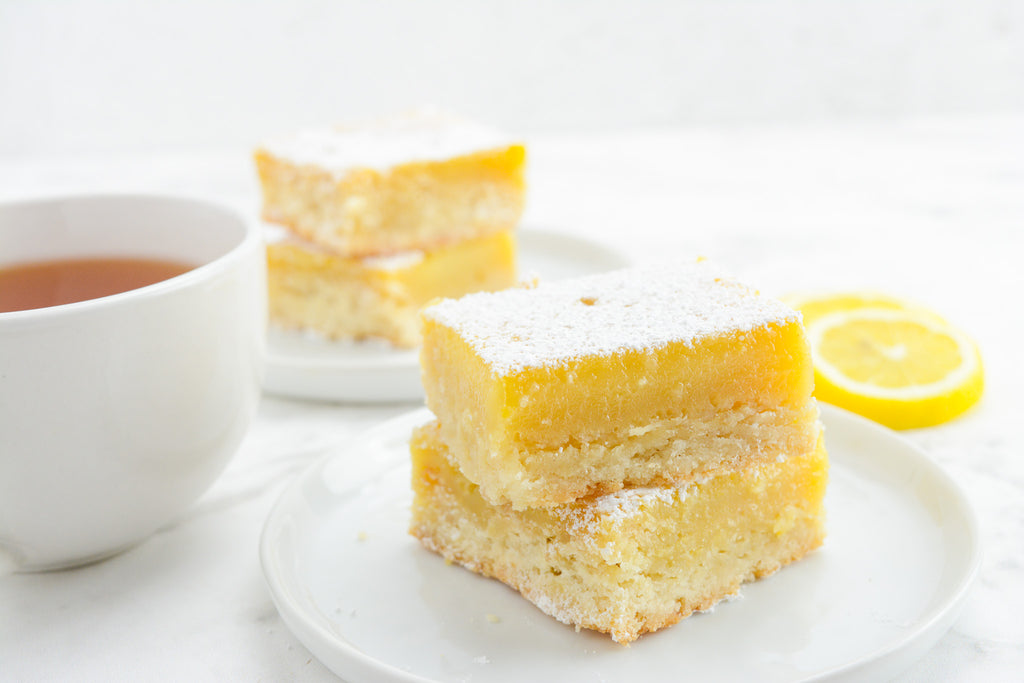White background with two small plates. Both plates have two lemon bars stacked. Cup of tea on the left side and two lemon slices on the right. 