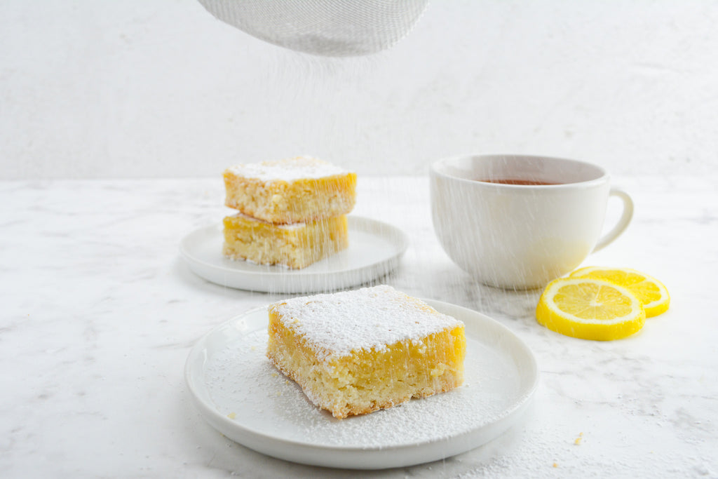 Two white plates sit on a white table with a cup of tea and lemon slices. A sifter sprinkles powdered sugar over the lemon bar.