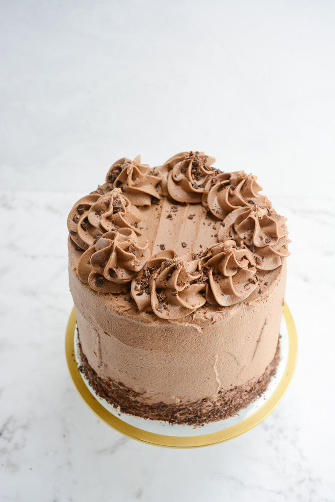 Tall light brown cake on a white background sits atop of a clear cake stand with gold trim. Decorated with chocolate shavings at the base and on the top with swirls of frosting.