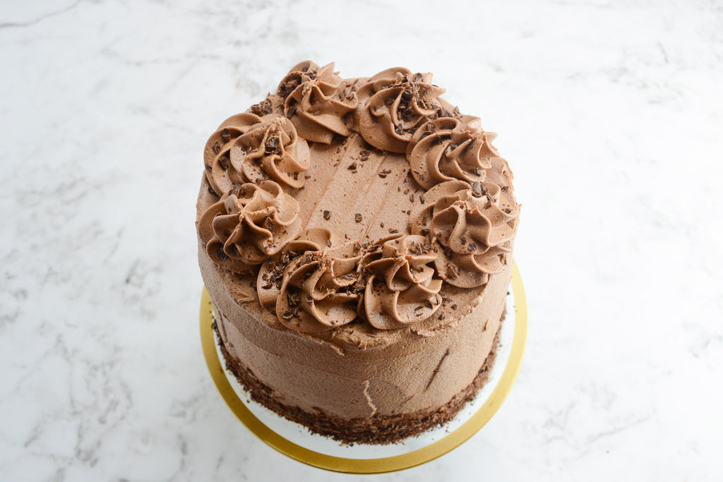 White marble background with a light brown cake sitting on top of a cake plate with a golden edge. Overhead view and swirls of frosting and chocolate shavings 