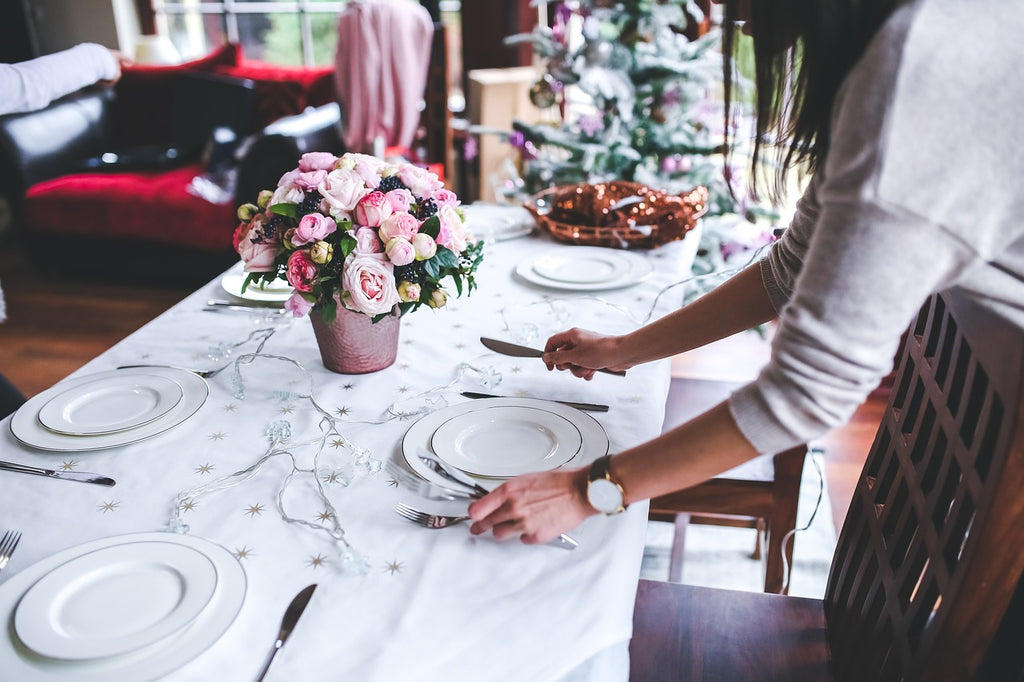 Person sets table with white plates and silverware. Pink flowers in a vase on the table and Christmas in the background