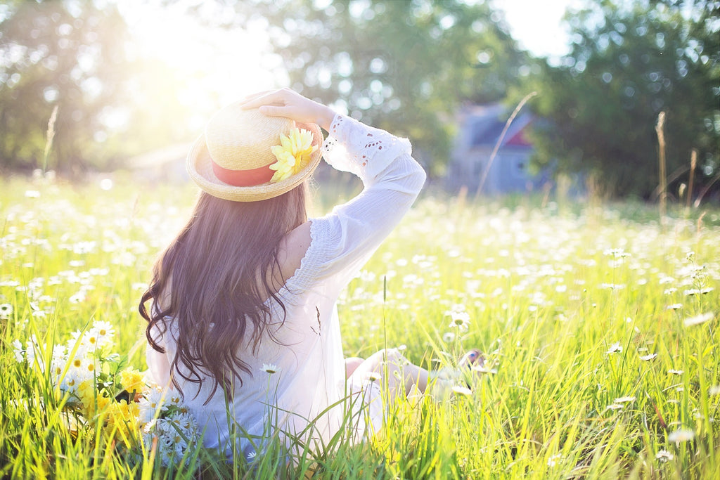 Woman in white shirt with long hair and tan hat sits in sun filled field with flowers