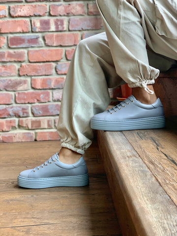 wide fitting grey leather trainers on model in front of brick background.