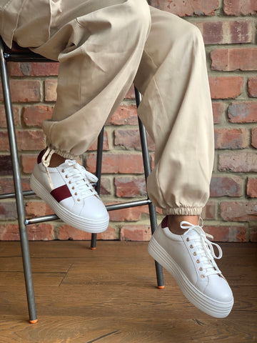 wide fitting white trainers with burgundy stripe in front of a brick background.