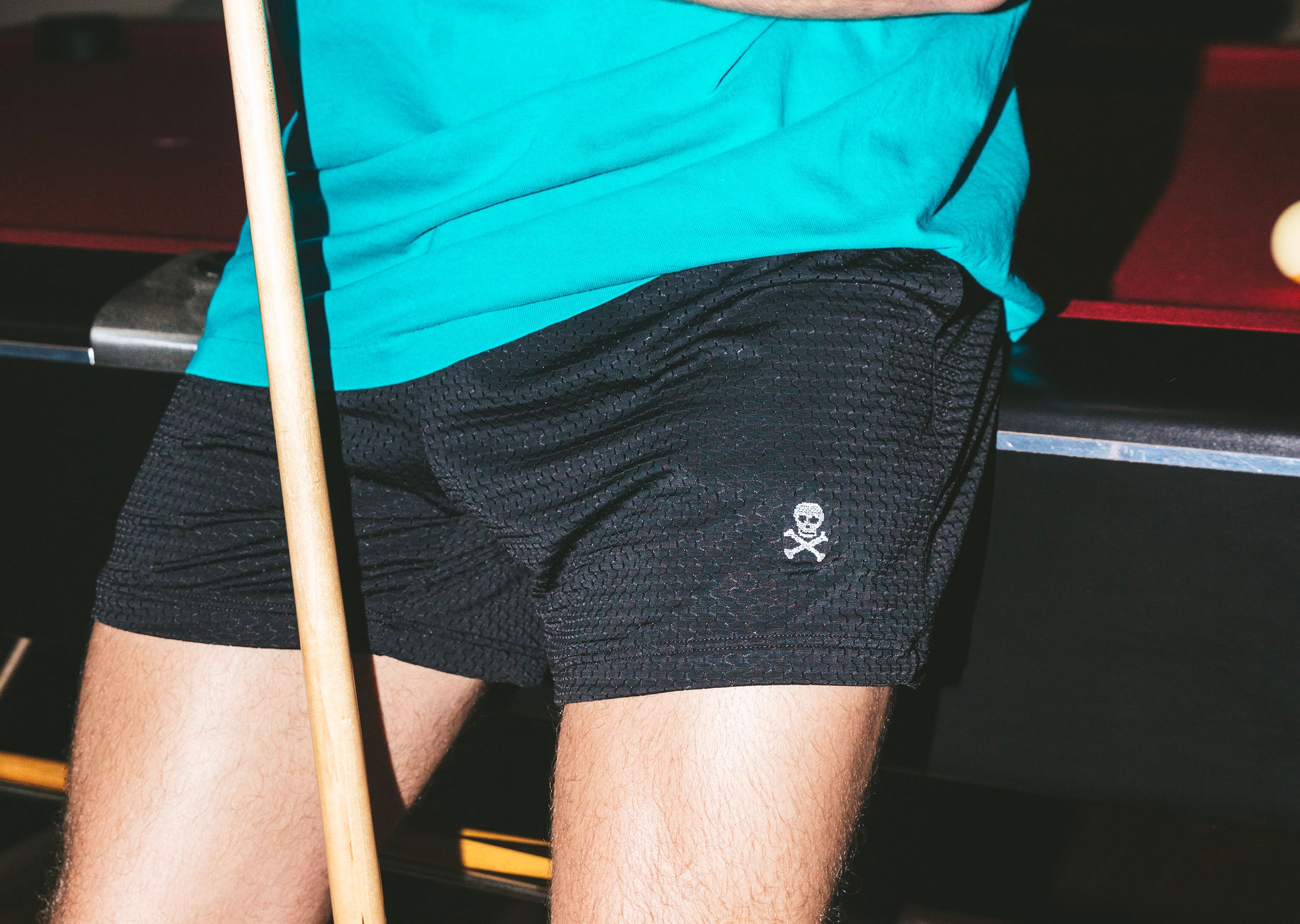 Man wearing black mesh shorts with white skull icon leaning against pool table.