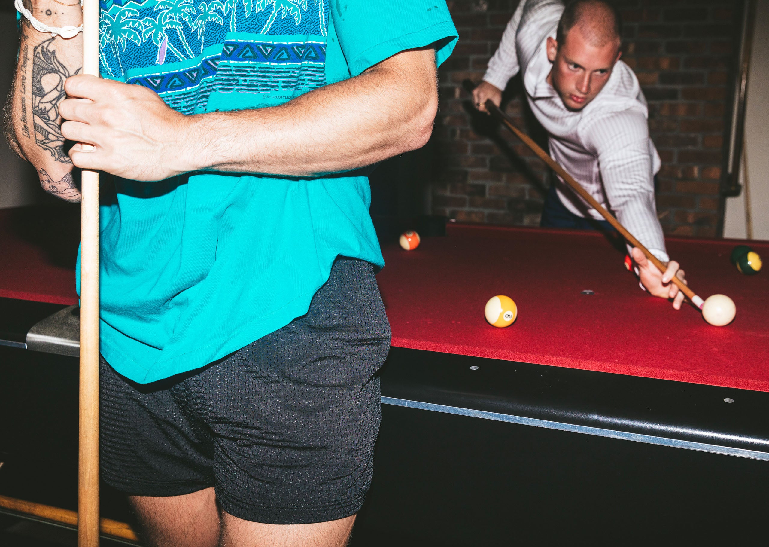 Two men playing pool with 1 close up wearing blue shirt and black mesh shorts.