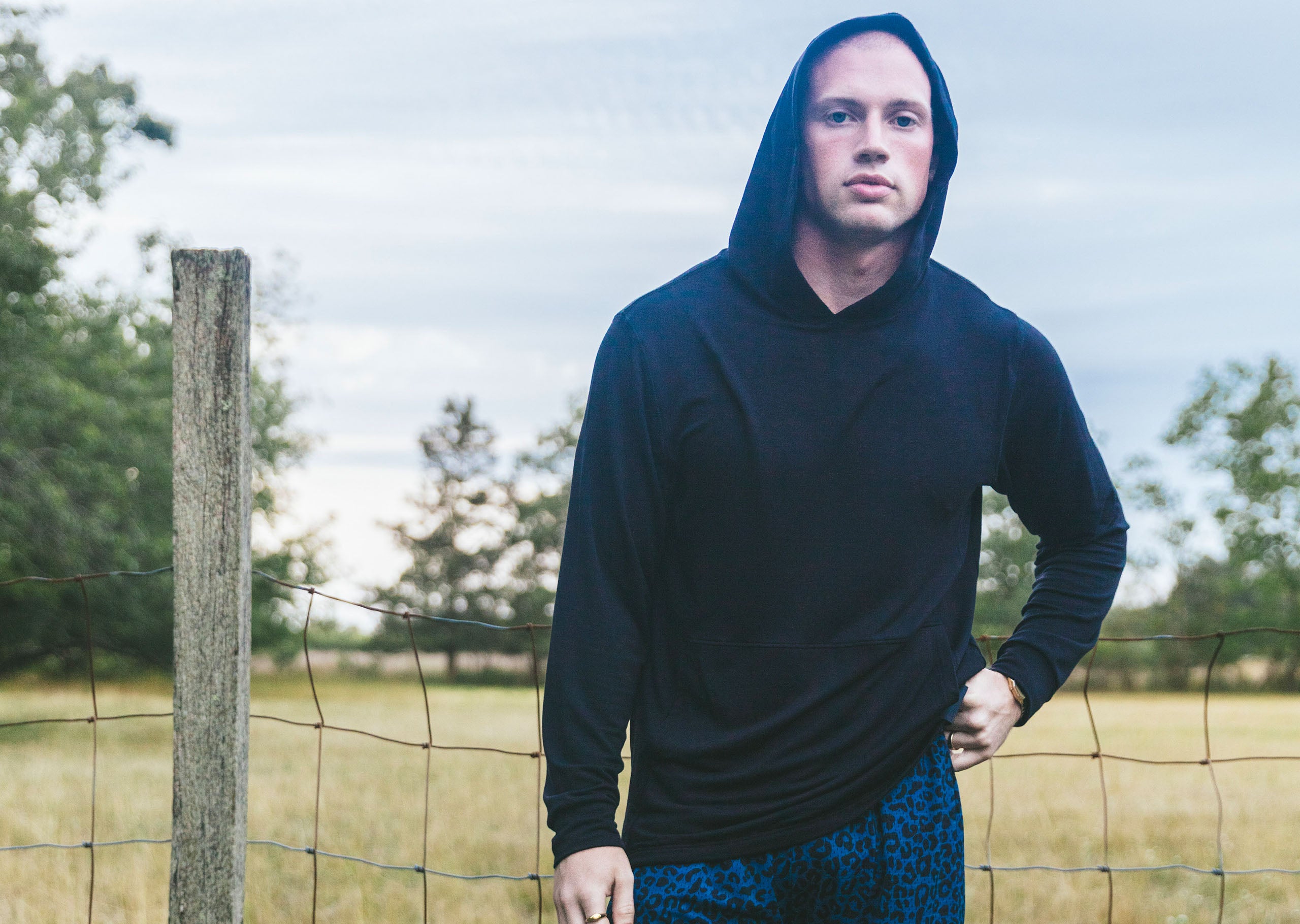 Man standing in field in front of fence wearing a black hoodie.