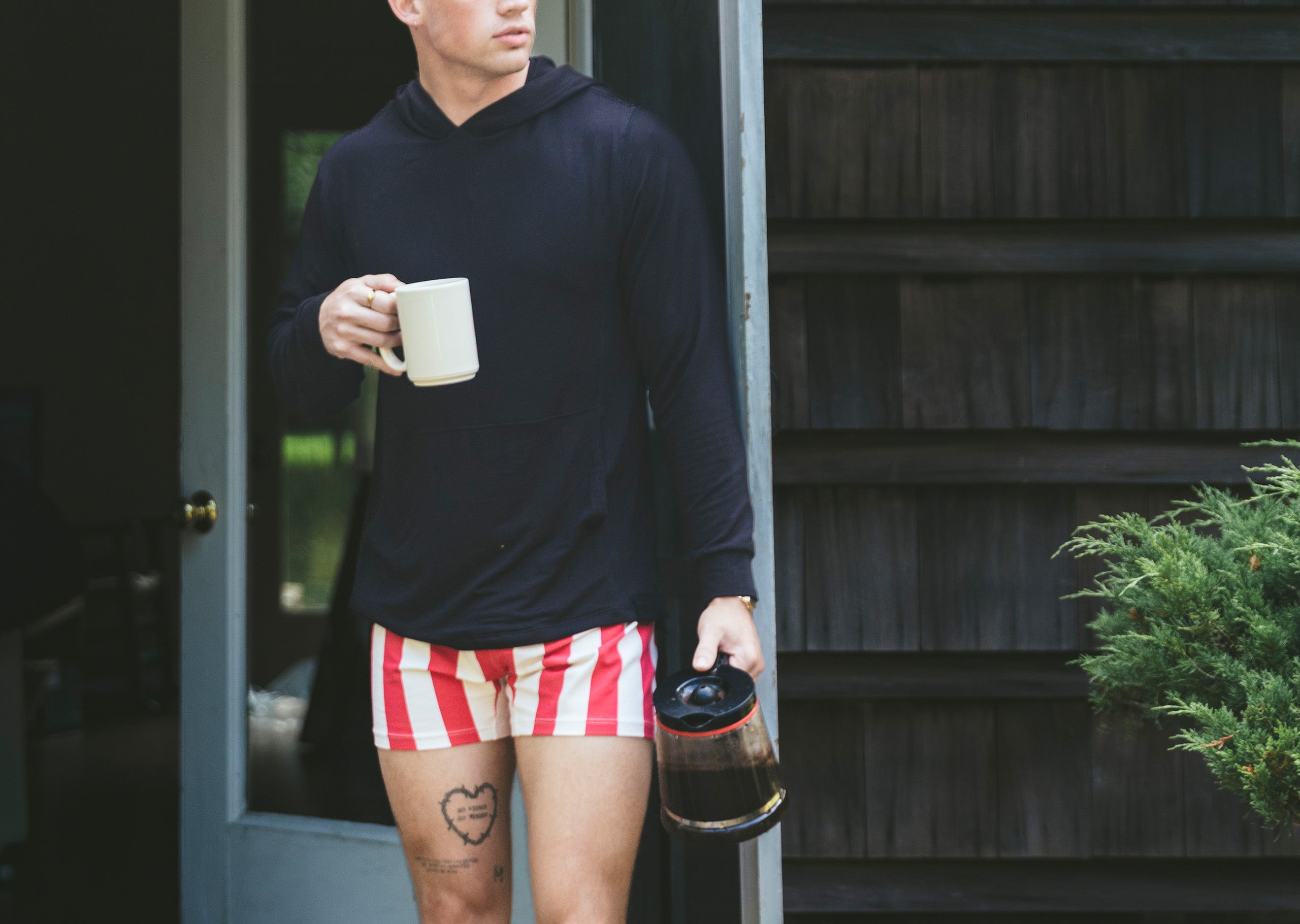 Man holding coffee pot in front of house wearing black sweat shirt with red and cream slim fit boxer.