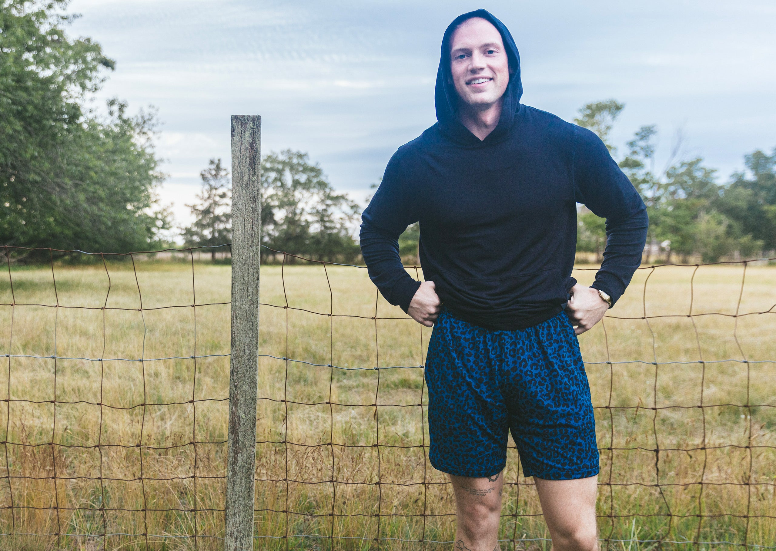 Man wearing black hoodie and midnight leopard printed lounge shorts standing next to fence in field.