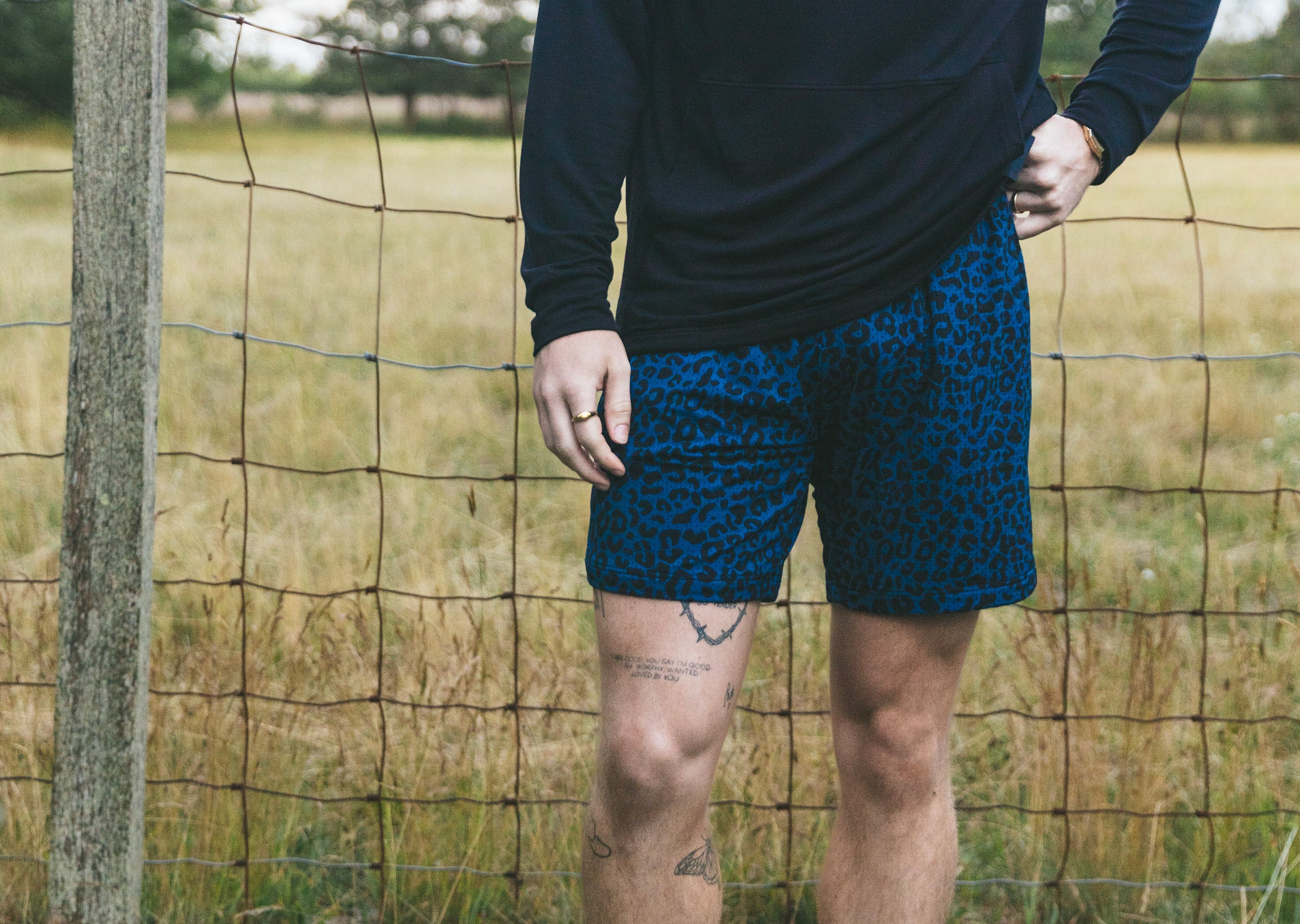 Man wearing black hoodie and midnight leopard lounge shorts standing by a fence in a field.