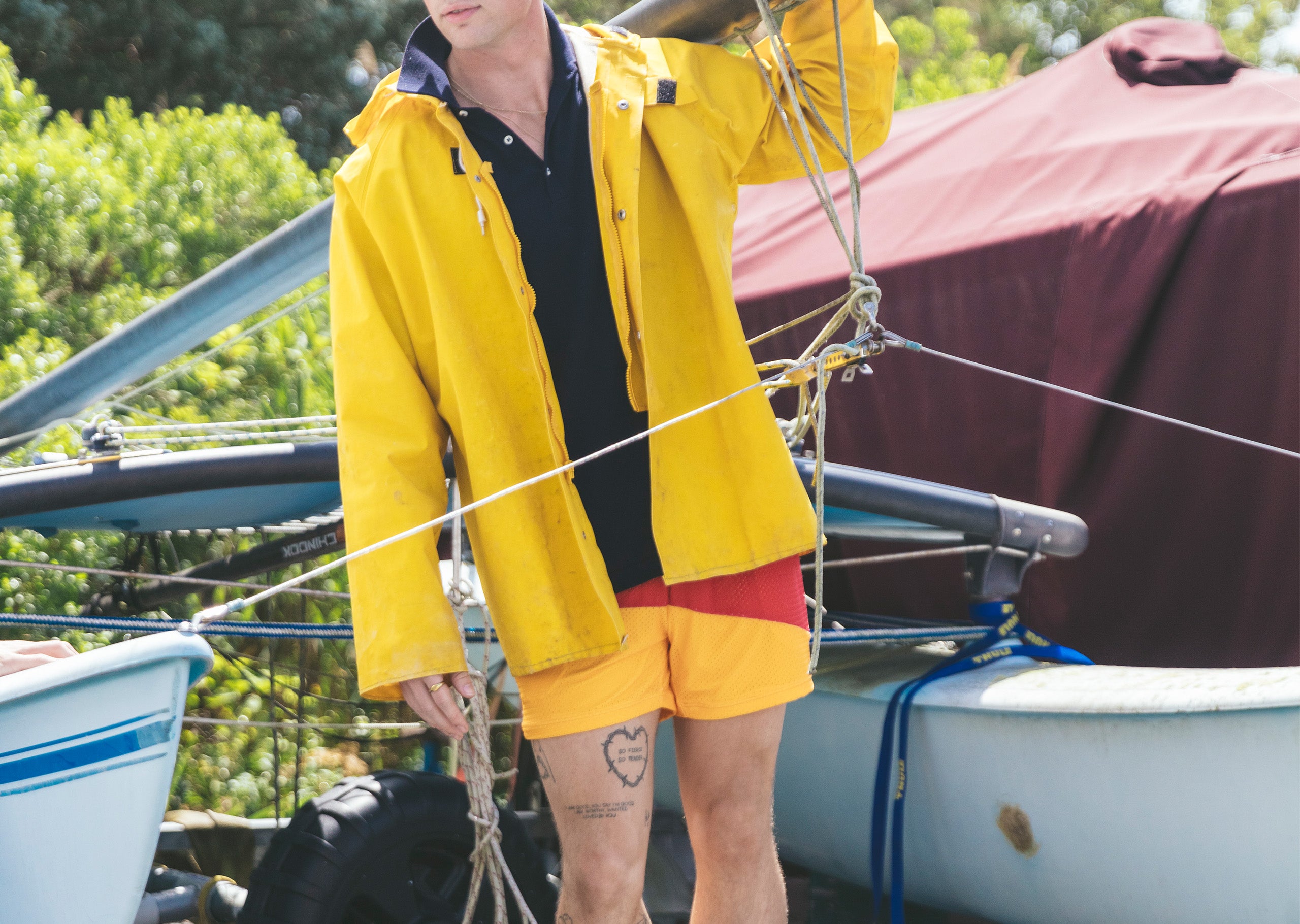 Man with yellow jacket and flipflops standing on a boat rack wearing Sunfish (red, blue and mustard) mesh lounge shorts.