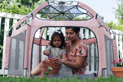 A mother is teaching her daughter to pot flowers while hanging out in the pop n go playpen