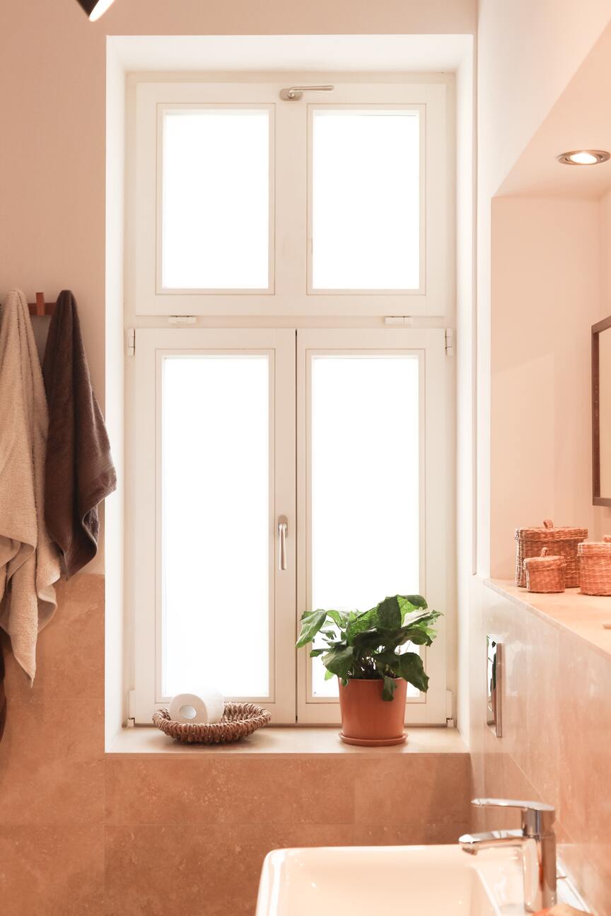 Pastellfarbenes Badezimmer mit Terrakottatopf auf dem Fenstersims, darin eine buschige, grüne Pflanze