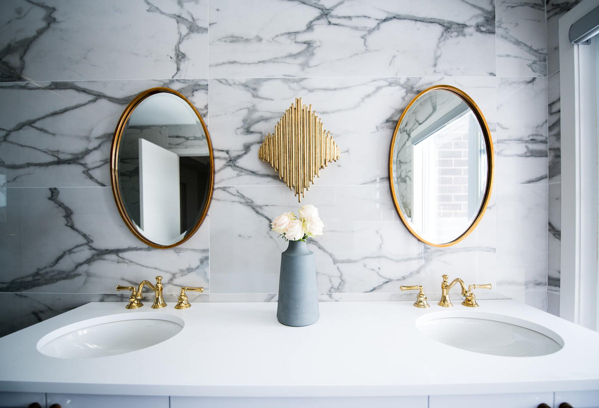 Marmorfarbene Wand im Badezimmer, doppeltes Waschbecken, zwei ovale Spiegel und in der Mitte ein Strauss weisser, kugelförmiger Trockenblumen