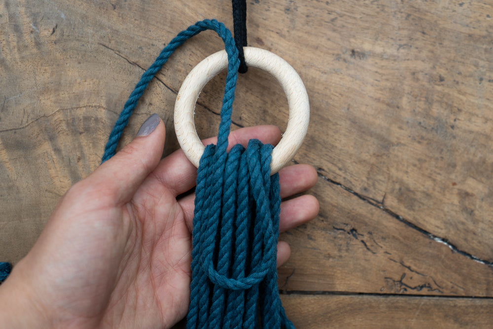 Bild für die Anleitung eines Wickelknoten mit Ring. Die Schnüre sind türkis und liegen auf einem Holztisch.