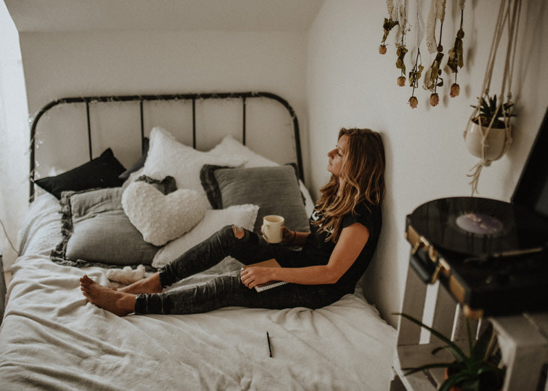 Blonde Frau sitzt mit einer Tasse Kaffee auf einem Bett mit vielen Kissen, hinter ihr diverse Pflanzen und eine grössere Sukkulente in Keramiktöpfen in Makramee-Hängern an der Wand