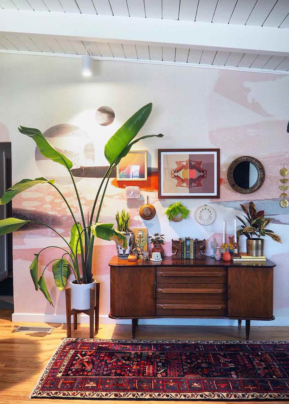 Eine XXL-Strelitzie in einem kleinen Holzständer neben einem hölzernen Sideboard mit weiteren Pflanzen und Deko-Artikeln, davor ein Teppich und an der Wand dahinter diverse viereckige und runde Bilder und Spiegel
