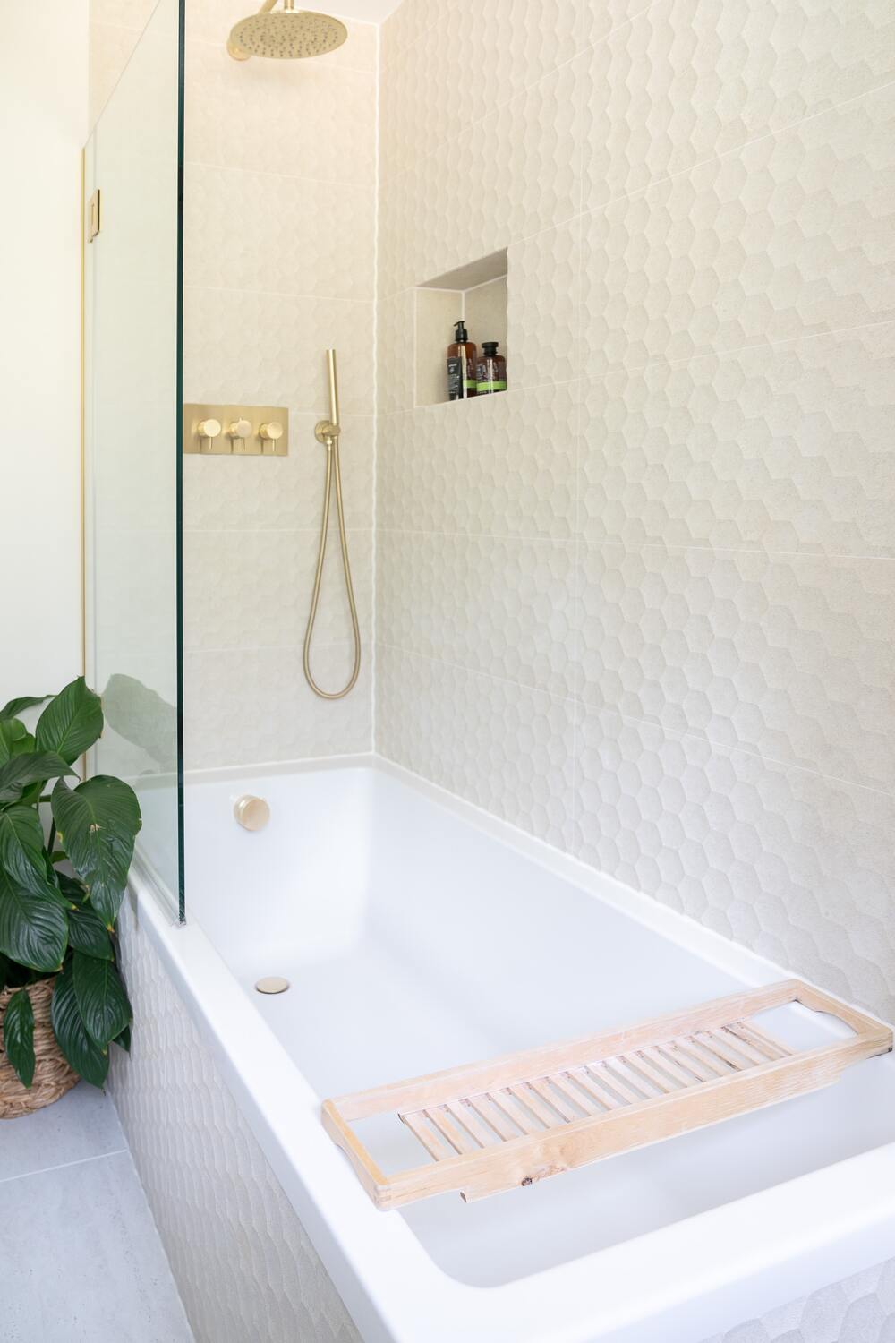 Eierschalenfarbene Wände, weisse Badewanne und goldene Armaturen, neben der Badewannendusche eine buschige Pflanze