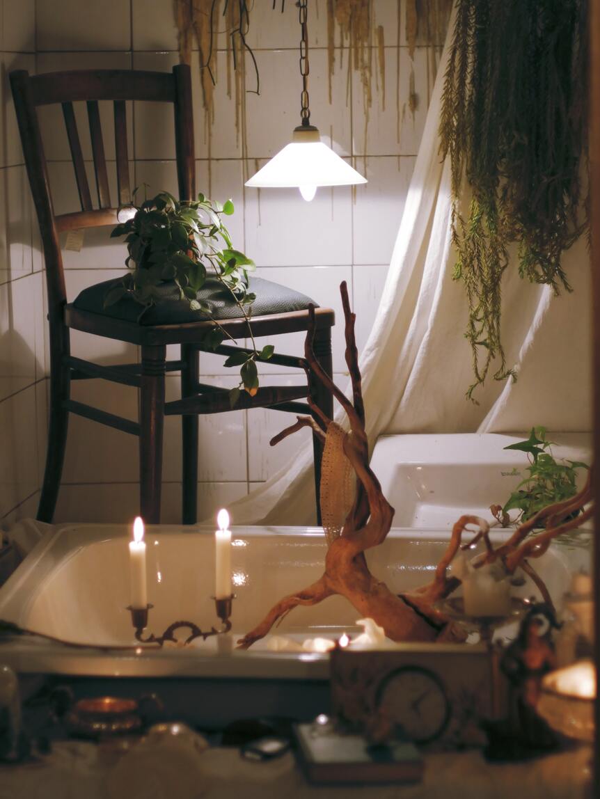 Mit Kerzenlicht beleuchtetes Badezimmer mit einer Hängepflanze auf dem Stuhl, einer an der Decke und diversen Dekoartikeln im Vordergrund
