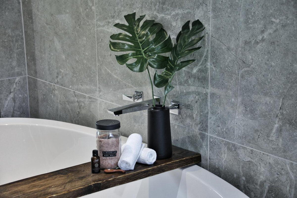 Grau marmorierte Wände, weisse Badewanne und auf einem Holzbrett darauf zwei Monstera-Stecklinge
