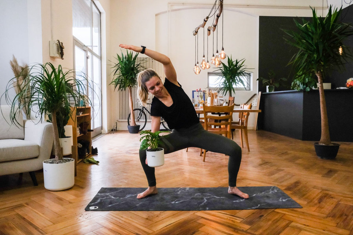 Plant-Yogi Hanna steht mit gebeugten Knien auf die rechte Seite geneigt da, in der rechten Hand, die auf dem rechten Oberschenkel ruht, die feey-Pflanze