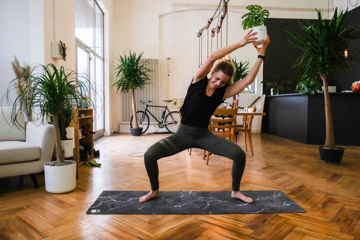 Plant-Yogi Hanna steht mit gebeugten Knien auf die linke Seite geneigt da, beide Arme mit der feey-Pflanze ausgestreckt