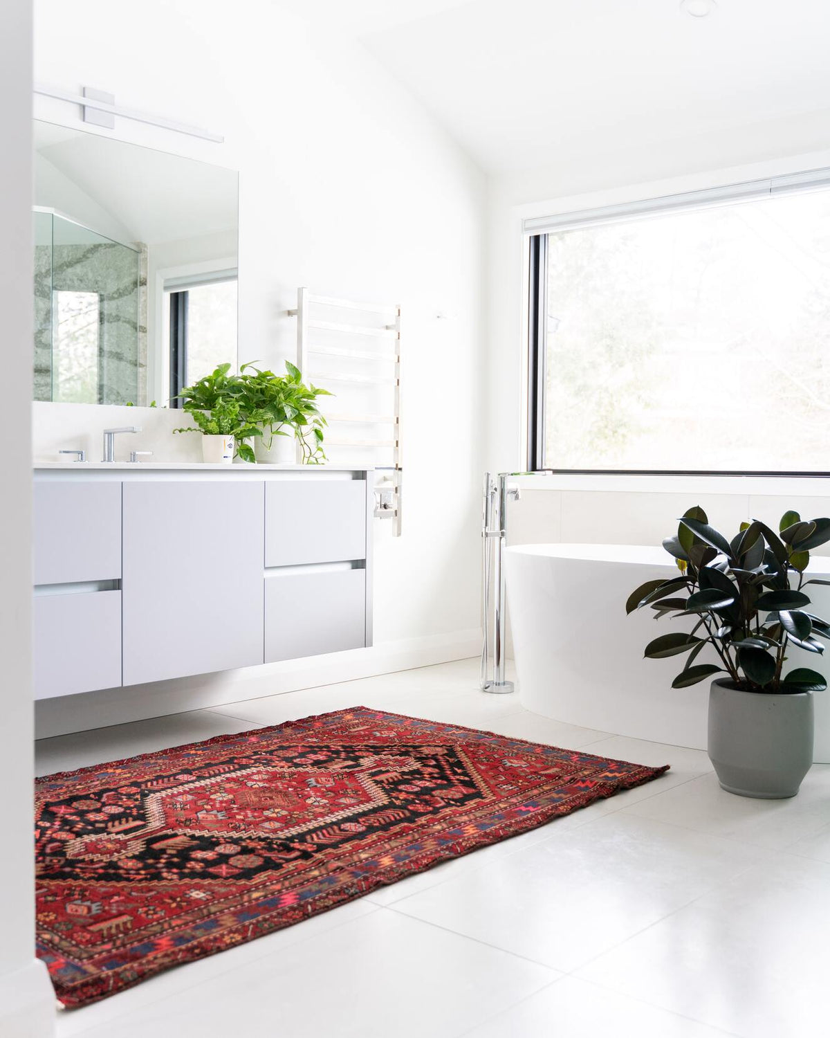 Helles Badezimmer mit einem orientalischen Teppich vor dem Waschbecken, auf letzerem Efeutute und Nestfarn, neben der Badewanne ein Gummibaum ein grauem Topf