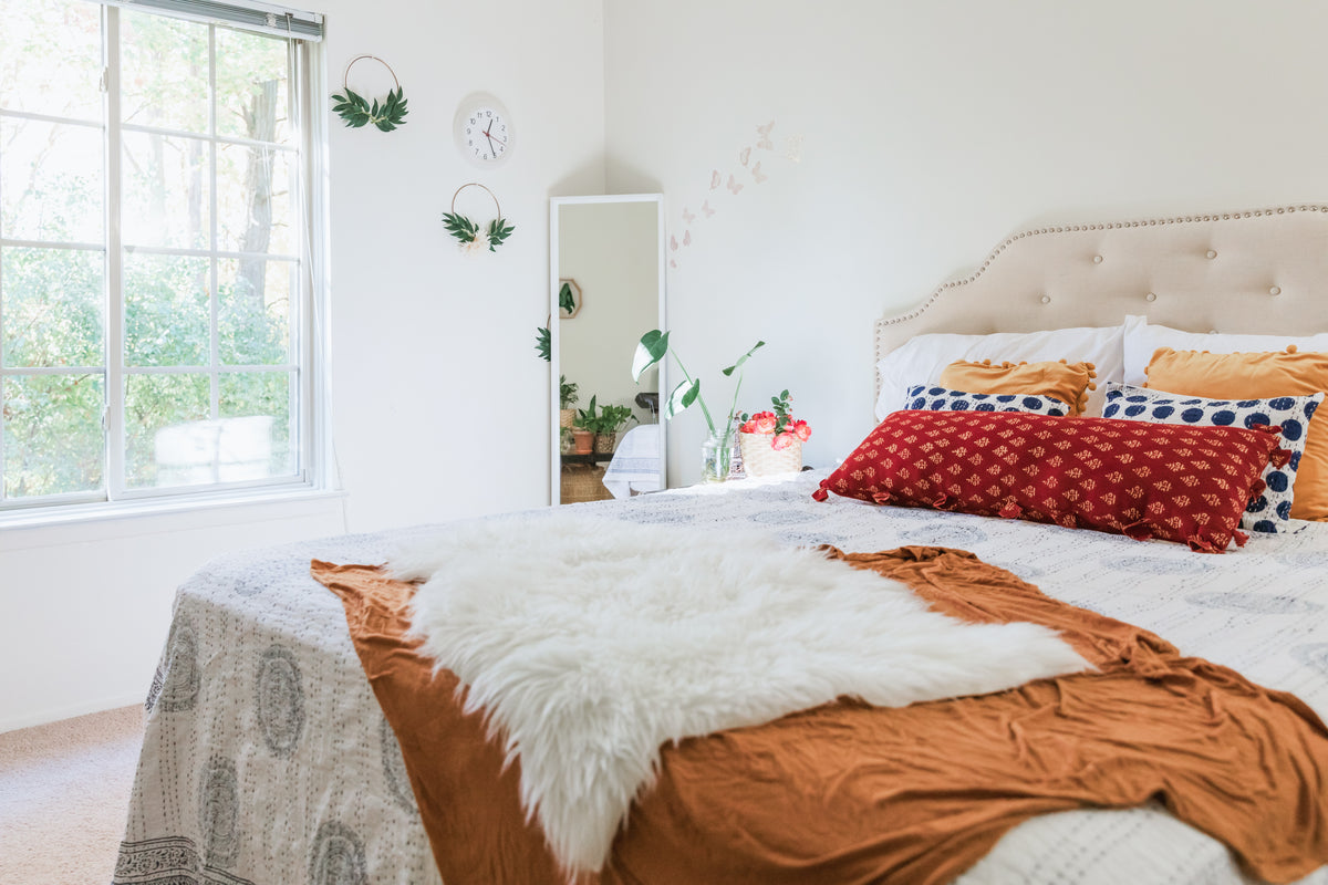 Auf einem cremefarbenen Bett liegen organge Kissen und eine orange Decke. In der Ecke des Zimmers hat es einen Spiegel und Pflanzen.