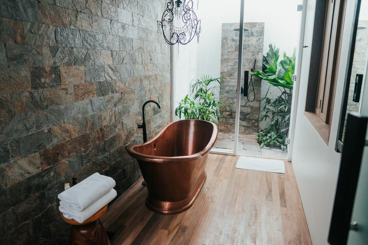 Luxuriöses Bad mit Holzboden, Steinwand, Messing-Badewanne und offener Dusche, darin Pflanzen
