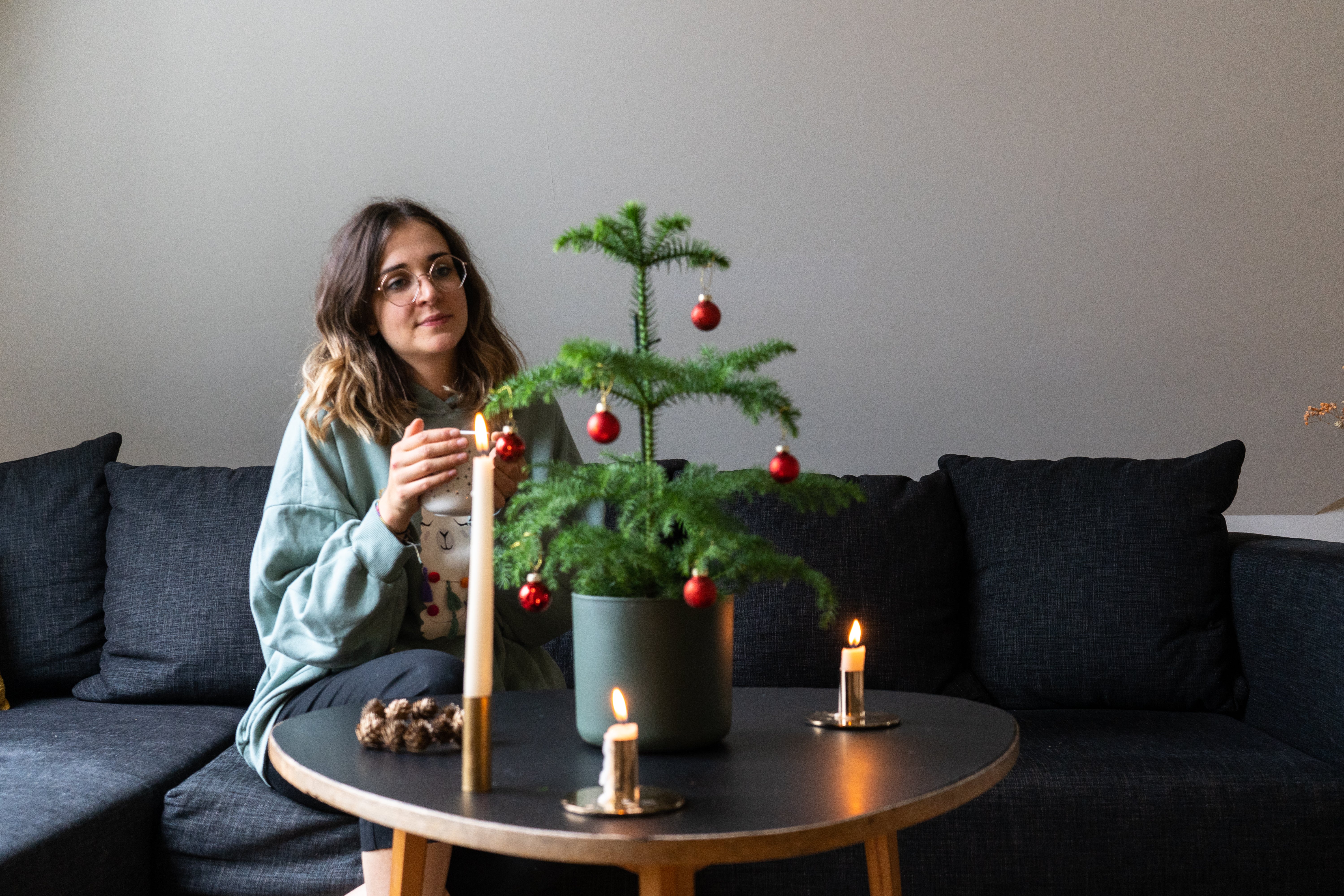 Frau mit braunen Haaren sitzt mit einer Tasse in der Hand auf einem Sofa, davor stehen ein geschmücktes Weihnachtsbäumchen und Kerzen