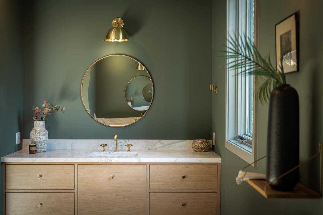 Dunkelgrüne Wand im Bad, daran ein runder Spiegel und davor das Waschbecken mit Marmor- und Holzverkleidung, im Vordergrund auf einem kleinen Wandvorsprung ein Pflanzentrieb in einer dunklen Vase