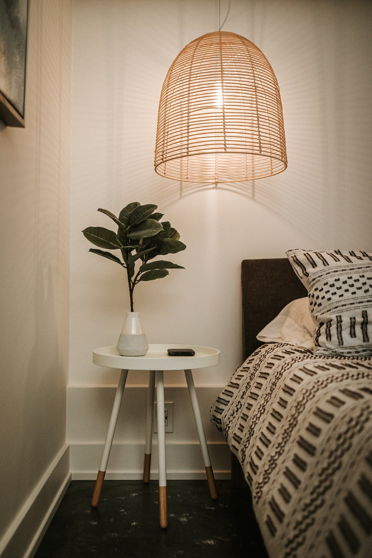 Ein Nachttisch steht neben einem Bett. An der Decke hängt eine grosse Lampe aus Bast. Auf dem Nachttisch steht eine Pflanze.