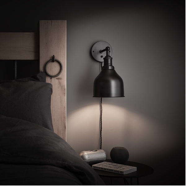 A black cone wall light shining over a bedroom side table