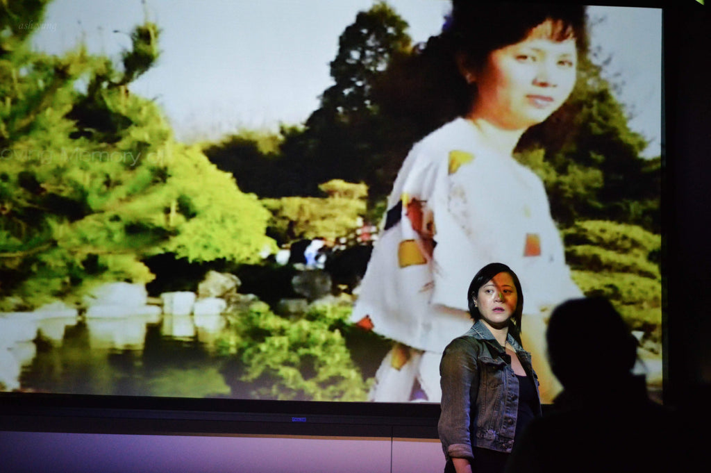 Susan Lieu standing in front of a screen projection of an old photo of her mother on stage during her performance.