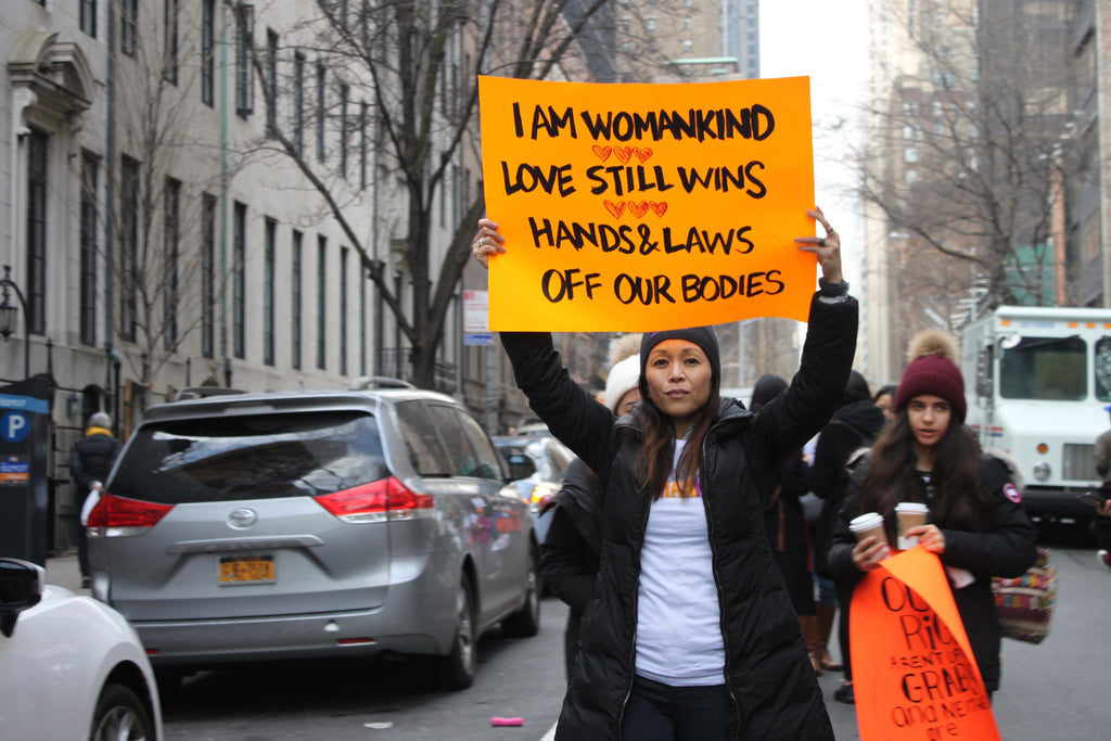 Woman protesting holds up an orange sign that reads "I am Womankind. Love still wins. Hands & laws off our bodies."