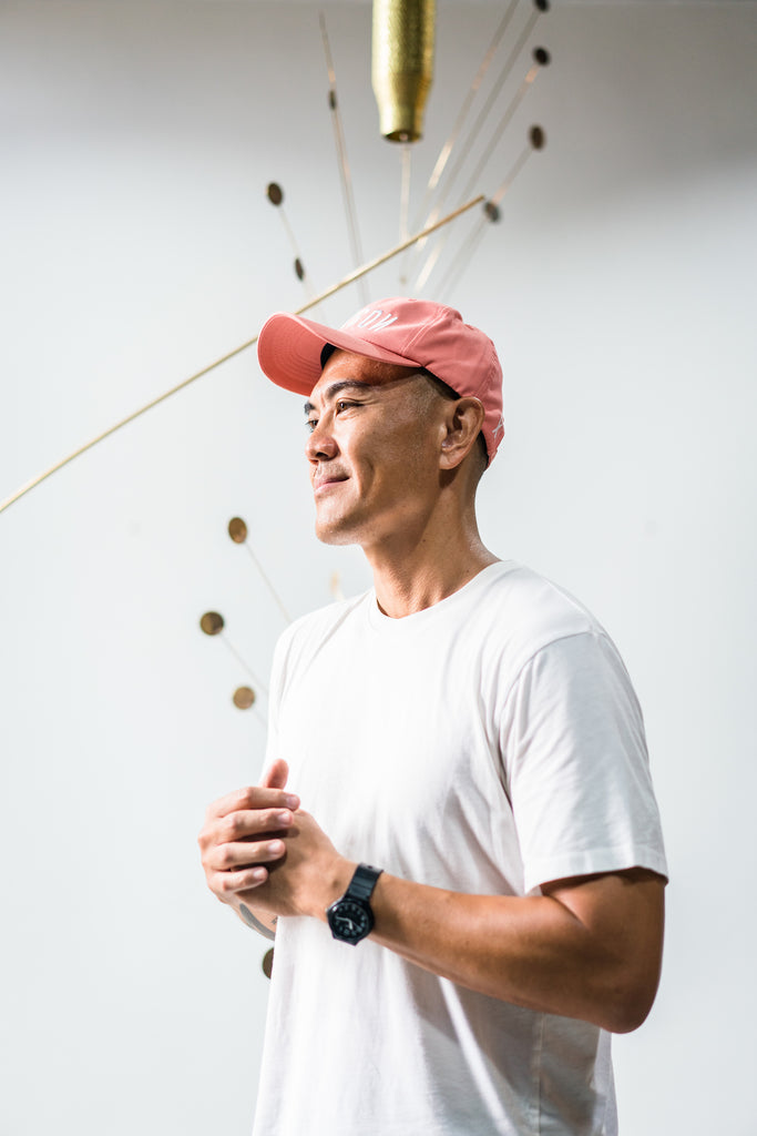 Tuan Andrew Nguyen in a white t-shirt, black watch, and red cap standing in front of his mobile artwork made from artillery shells.