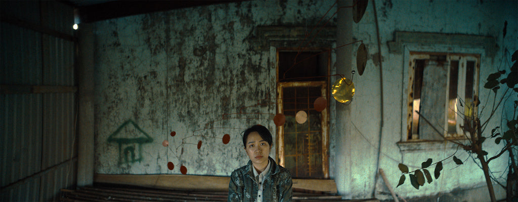A young woman sits in front of her home with mobiles hanging around her. She is the main character of the film "The Unburied Sounds of a Trouble Horizon."