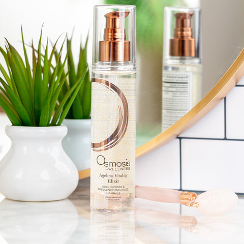 Osmosis Ageless Vitality Elixir on a counter next to a copper rimmed mirror with a green plant in a white vase