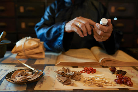 Person sitting at table with instruments and ingredients of traditional Chinese medicine. Only their hands are visible as they hold a bottle