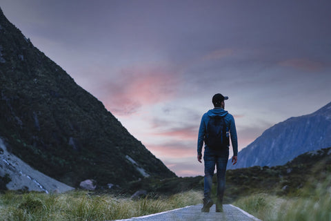 A silhouette of a man on a hike.  He's looking out toward a purple/blue sky and mountains.