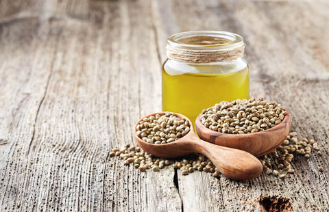 A glass bottle of hemp seed oil on a wooden table, with a wooden spoon holding hemp seeds