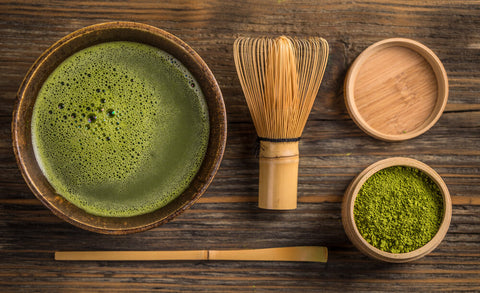 A bird's eye view of matcha tea on a wooden bench. There's a bowl of powder, a cup of tea and a whisk.