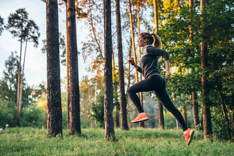 A woman running outdoors, she's wearing black with pink runners. The grass is green and lush with trees around.