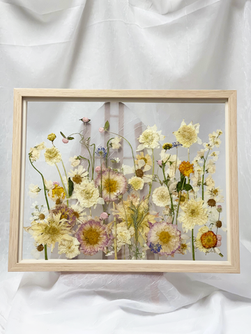 Large pressed flower frame designed in a field like design on an easel against a white linen backdrop.
