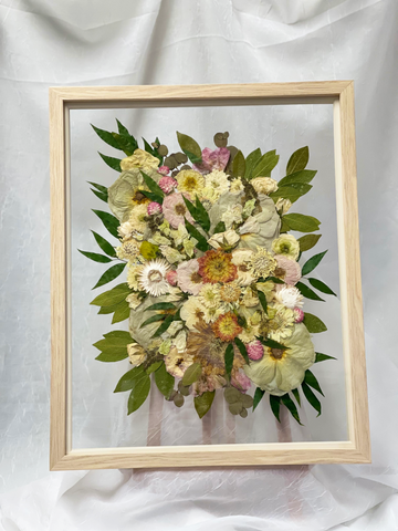 Pressed flowers encased in a large frame on an easel against a white linen backdrop.
