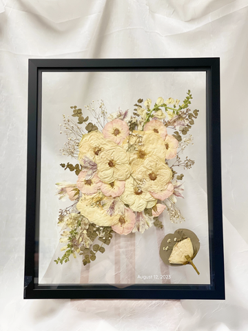 Large black frame with pressed flowers and a date on it with a boutonniere in the corner against a white background.