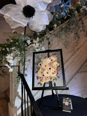 pink and white pressed flowers encased in a black frame on an easel on front of a staircase.