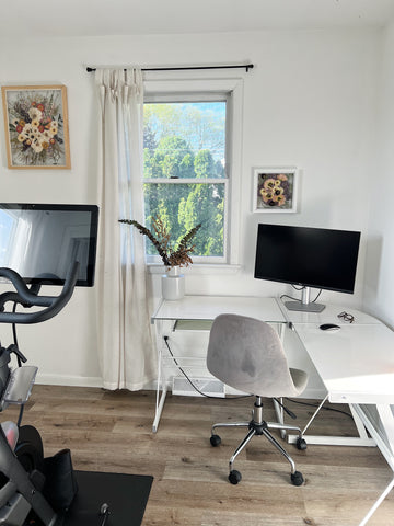 Home office with a desk, computer, and exercise bike. Two pressed flower frames are hanging on either side of the window.
