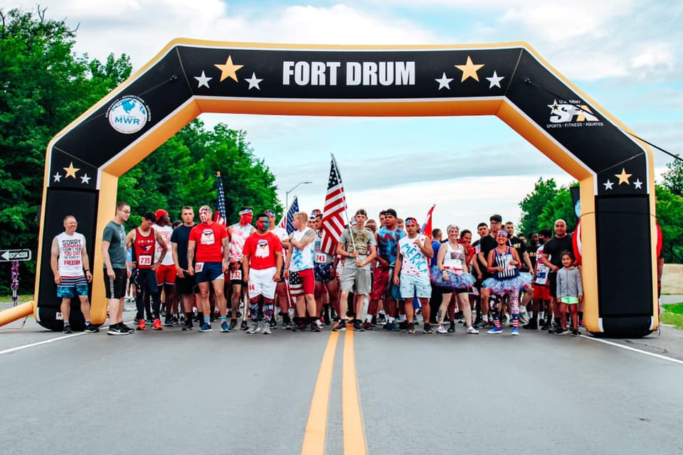 A spirited group gathers at the starting line under a Fort Drum branded denier polyester inflatable arch, ready to embark on their race.