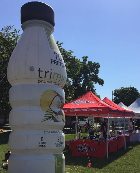 Large inflatable bottle of 'trimino' protein-infused coconut water displaying nutritional information, with event tents including one for 'Boston Cannons' in the background.